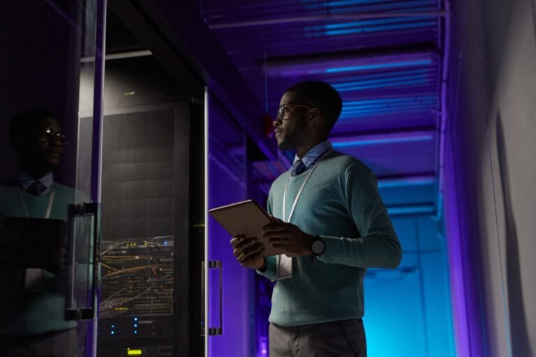 A young African American IT specialist holding a tablet while inspecting server racks in a dimly lit data center with blue and purple lighting.