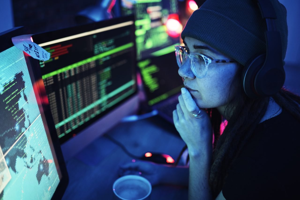 A woman wearing headphones intensely focusing on computer screens displaying programming and cybersecurity tasks